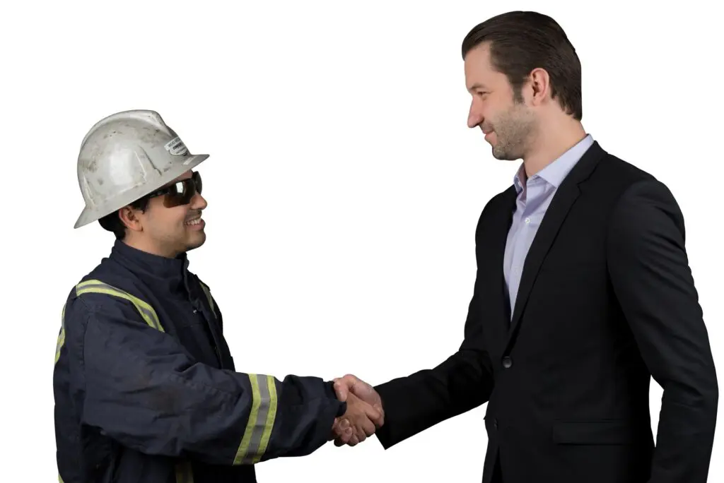 A man in a dark suit shakes hands with a smiling worker in a hard hat and safety jacket