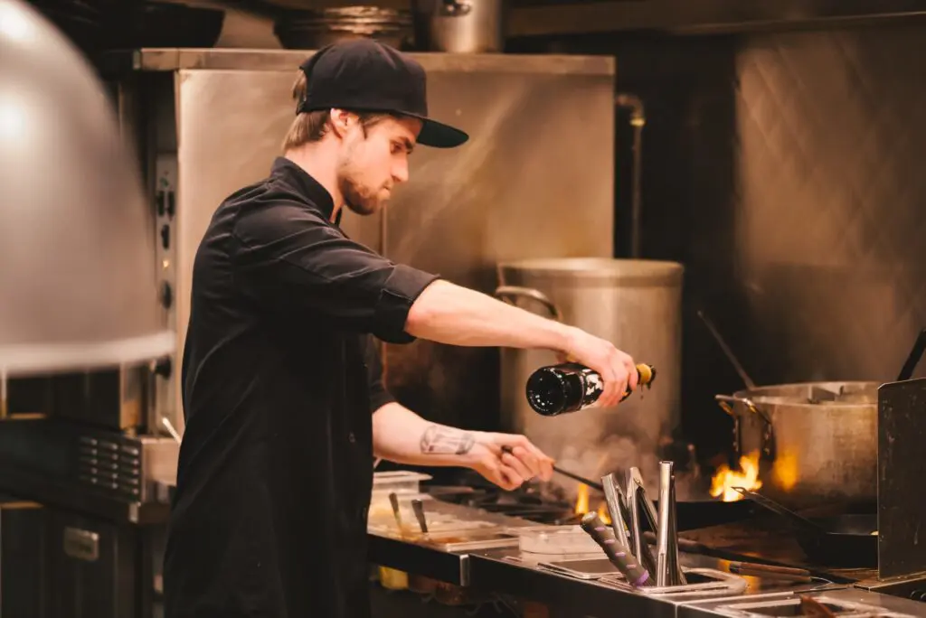 A chef in a black cap pours wine into a pan over open flame in a busy restaurant kitchen