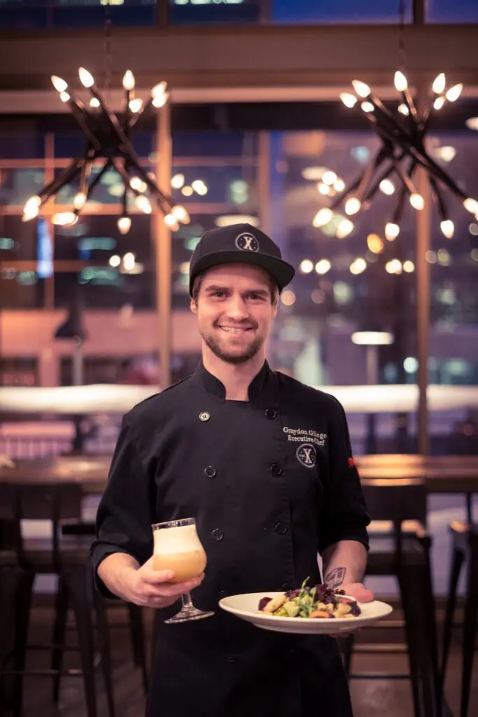 A smiling chef in a black jacket and cap holds a plated dish and a beer glass in a restaurant