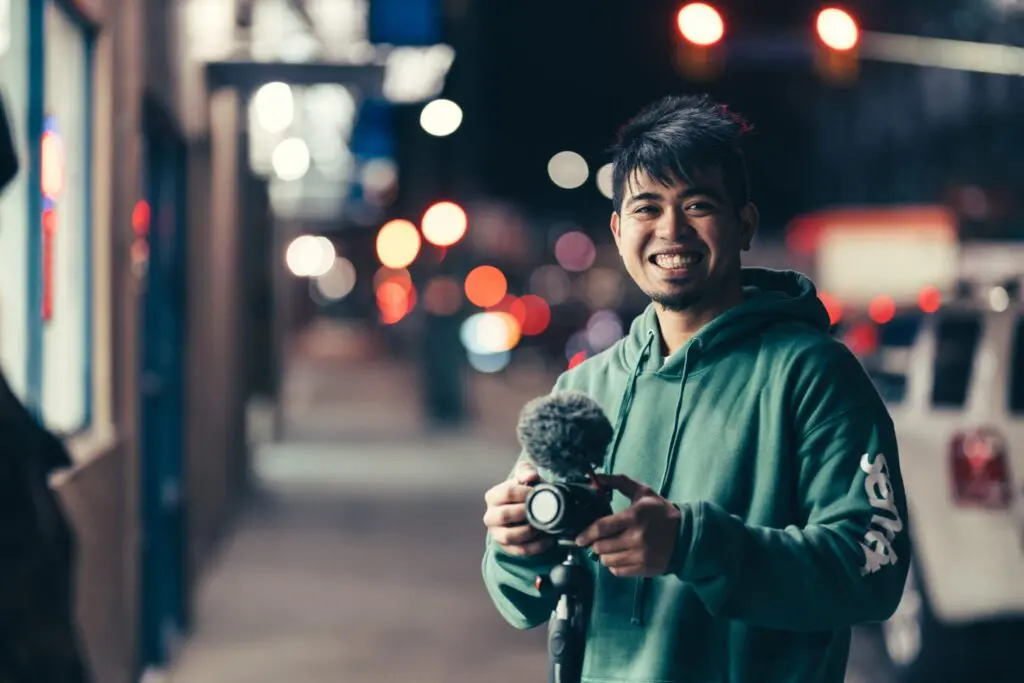 Smiling man in a green hoodie holding a camera on a Kelowna street at night