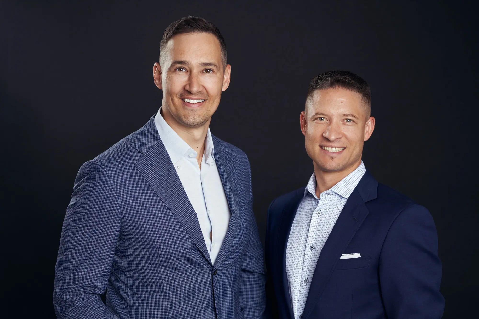 Two men in blazers smiling together in a studio headshot on a dark backdrop
