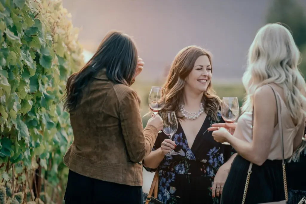 Three women laughing and holding wine glasses beside grapevines at sunset on an Okanagan wine tour with Wicked Wine Tours
